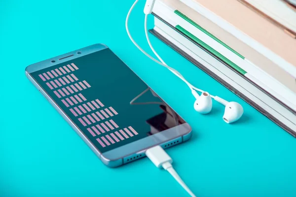 Teléfono con auriculares blancos junto a una pila de libros sobre un fondo azul. Concepto de audiolibros y educación moderna — Foto de Stock