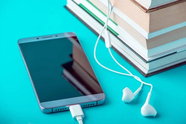 Phone with white earphones next to a stack of books on a blue background. Concept of audiobooks and modern education