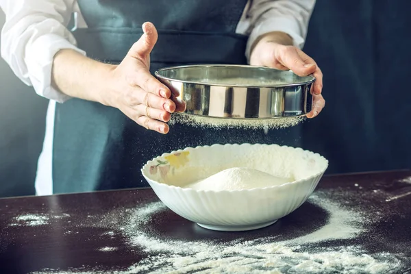 The chef in black apron sifts the flour through a sieve to prepare the dough for pizza — Stock Photo, Image