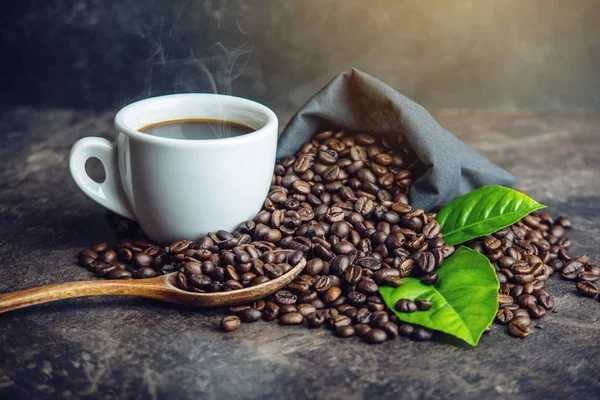 Taza de café negro blanco con un montón de granos de café y hojas verdes en el bolso sobre fondo oscuro —  Fotos de Stock