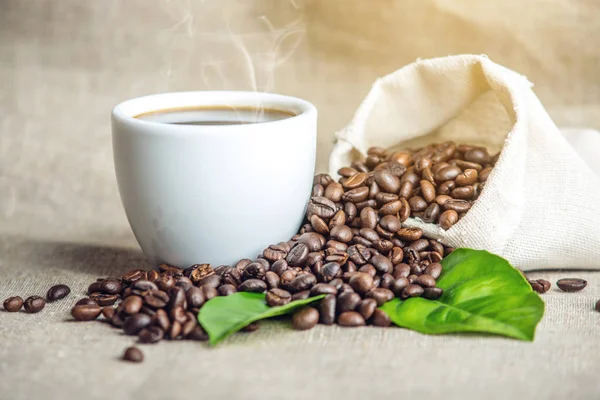 Taza de café espresso negro blanco con una pila de granos de café y hojas verdes en bolsa sobre fondo de lino blanco —  Fotos de Stock