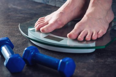 Kadın ayakları koyu arka plan üzerinde dumbbells ile kilo kontrolü için elektronik teraziler tutuyoruz. Spor eğitim, beslenme ve kilo kaybı kavramı