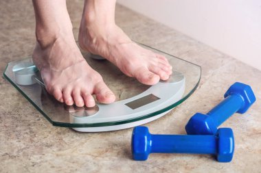 Kadın ayakları elektronik ayakta ölçekler üzerinde açık renkli dumbbells ile kilo kontrolü için. Spor eğitim, beslenme ve kilo kaybı kavramı