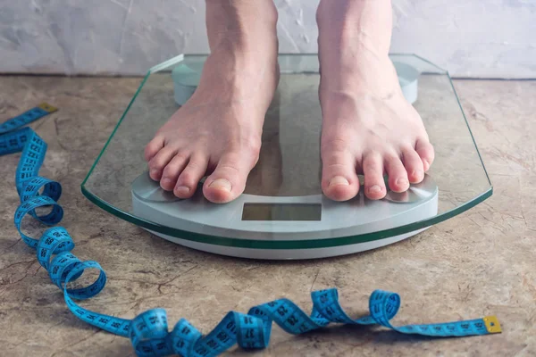 Female feet standing on electronic scales for weight control with and measuring tape on light background. The concept of sports training, diets and weight loss