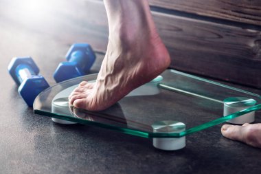 Kadın ayakları koyu arka plan üzerinde dumbbells ile kilo kontrolü için elektronik teraziler tutuyoruz. Spor eğitim, beslenme ve kilo kaybı kavramı