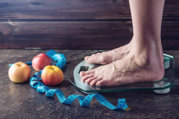 Female feet standing on electronic scales for weight control with measuring tape and apples on dark background. The concept of sports training, diets and weight loss