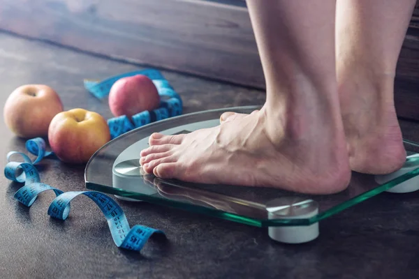 Female feet standing on electronic scales for weight control with measuring tape and apples on dark background. The concept of sports training, diets and weight loss
