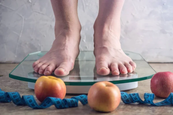 Female feet standing on electronic scales for weight control with measuring tape and apples on light background. The concept of sports training, diets and weight loss