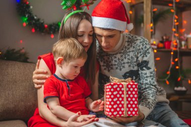 Anne ve baba oğullarına kırmızı bir kutuda hediye veriyorlar. Çocuklu genç bir aile Noel 'i ve yeni yılı evde kutluyor.