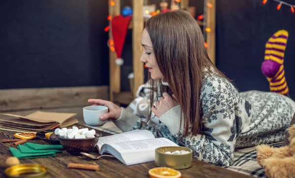 Giovane ragazza in maglione che legge un libro con una tazza di caffè la sera in una calda atmosfera natalizia. umore di anno nuovo — Foto Stock