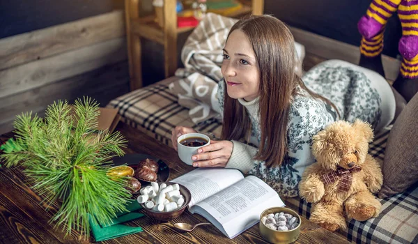 Giovane ragazza in maglione che legge un libro con una tazza di tè la sera in una calda atmosfera natalizia. umore di anno nuovo — Foto Stock