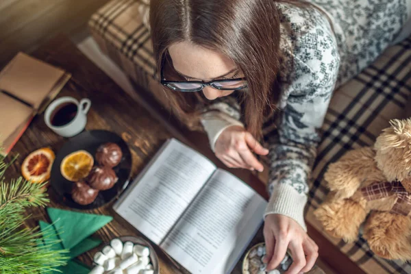 Ragazza in maglione che legge un libro con una tazza di caffè la sera in una calda atmosfera natalizia. umore di anno nuovo — Foto Stock