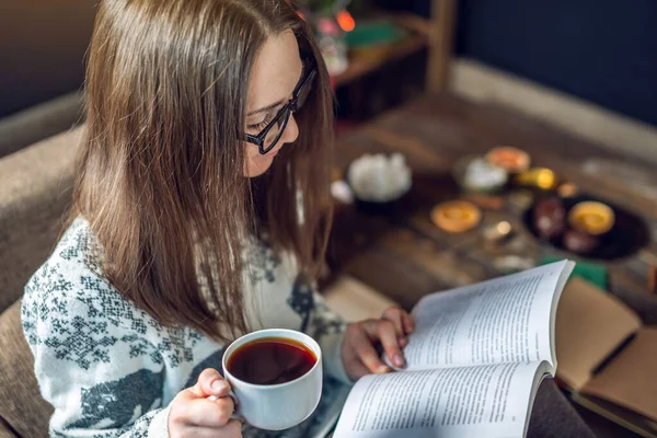 Giovane ragazza in maglione che legge un libro con una tazza di caffè la sera in una calda atmosfera natalizia. umore di anno nuovo — Foto Stock