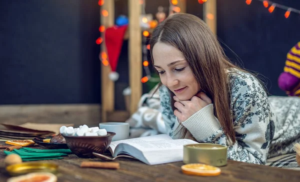 Jeune fille en pull lisant un livre avec une tasse de café le soir dans une ambiance chaleureuse de Noël. Nouvel an humeur — Photo