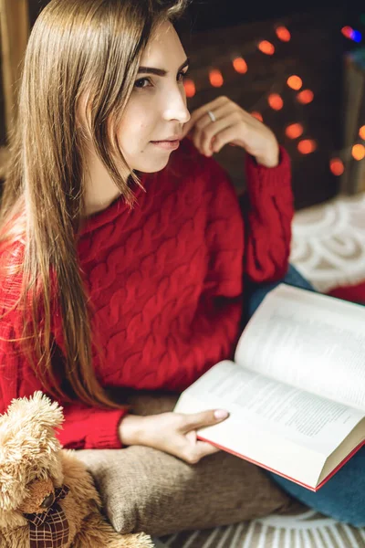 Egy piros pulóveres nő könyvet olvas esténként, meleg karácsonyi hangulatban. Hangulatos újévi hangulat — Stock Fotó