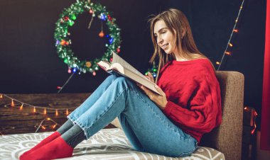 Sıcak bir Noel atmosferinde, kırmızı kazaklı bir kadın, arka planda ışık çelenkleriyle kitap okuyor.