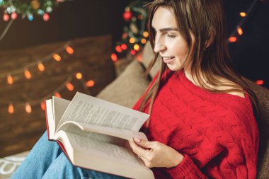 Sıcak bir Noel atmosferinde, kırmızı kazaklı bir kadın, arka planda ışık çelenkleriyle kitap okuyor.