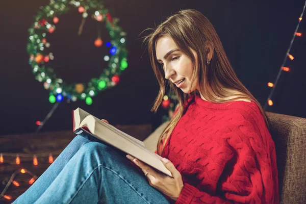 Woman in a red sweater reading a book in the evening in a warm Christmas atmosphere on background of lights garlands