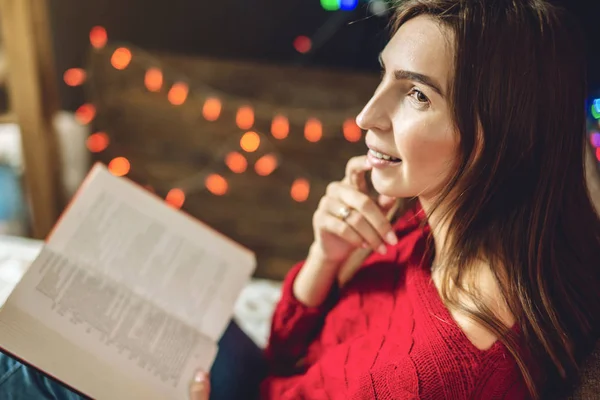 Femme en pull rouge lisant un livre le soir dans une ambiance chaleureuse de Noël sur fond de guirlandes de lumières — Photo