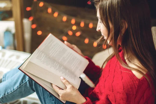 Femme en pull rouge lisant un livre le soir dans une ambiance chaleureuse de Noël sur fond de guirlandes de lumières — Photo