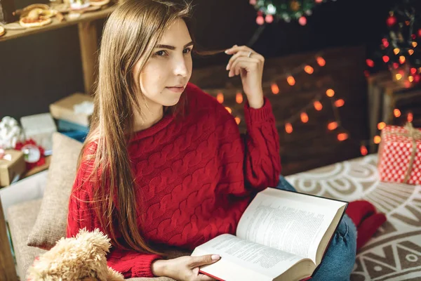 Femme en pull rouge lisant un livre le soir dans une ambiance chaleureuse de Noël. Confortable nouvelle humeur de l'année — Photo