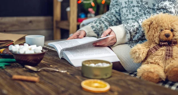 Fiatal lány pulóverben könyvet olvas egy bögre kávéval este, meleg karácsonyi hangulatban. Újévi hangulat — Stock Fotó