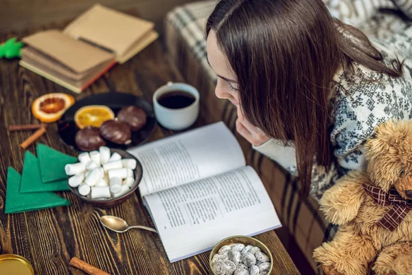 Jeune fille en pull lisant un livre avec une tasse de café le soir dans une ambiance chaleureuse de Noël. Nouvel an humeur — Photo