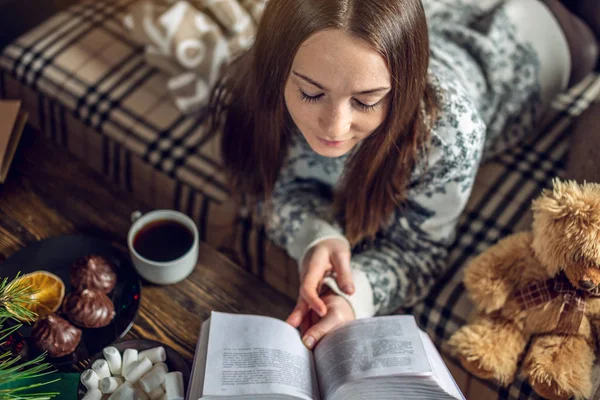 Fiatal lány pulóverben könyvet olvas egy bögre teával este, meleg karácsonyi hangulatban. Újévi hangulat — Stock Fotó