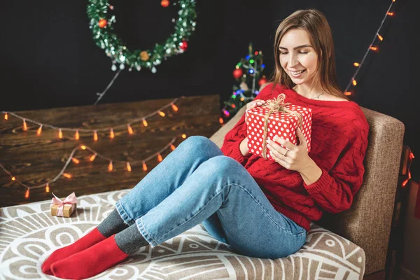 Wanita bahagia dengan sweater merah duduk di sofa dengan kotak hadiah di tangannya pada malam Natal. Suasana tahun baru yang nyaman Stok Gambar Bebas Royalti