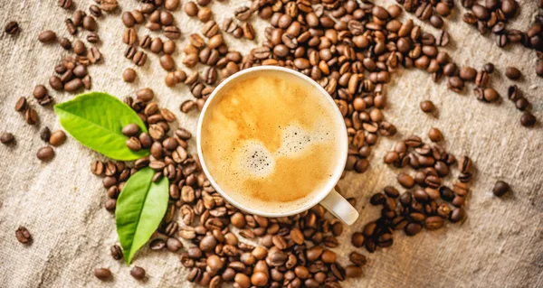 Tazza di caffè con schiuma sui grani sparsi sulla iuta. Fragrante bevanda tonificante di fagioli appena tostati. — Foto Stock