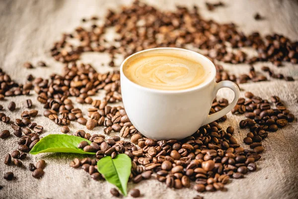 Tazza di caffè con schiuma sui grani sparsi sulla iuta. Fragrante bevanda tonificante di fagioli appena tostati. — Foto Stock