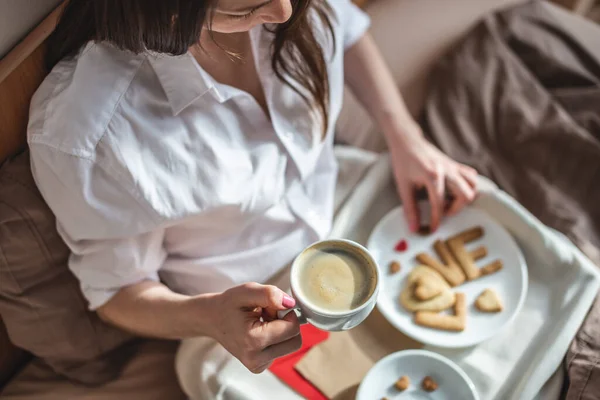 La mujer joven tiene un desayuno romántico en la cama de la mañana. Café y palabra amor de las galletas. Sorpresa en el día de San Valentín — Foto de Stock