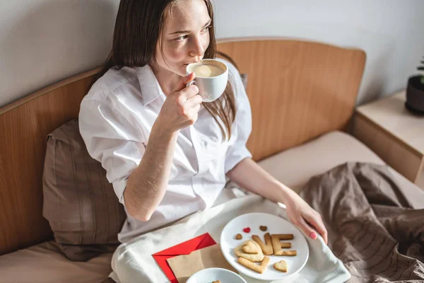 La mujer joven tiene un desayuno romántico en la cama de la mañana. Café y palabra amor de las galletas. Sorpresa en el día de San Valentín — Foto de Stock