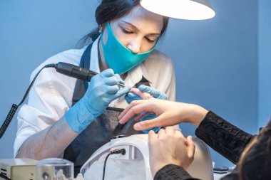 Salondaki bir kadın manikür ustası müşterinin tırnaklarıyla mavi arka planda çalışıyor. Kozmetik ve tırnak bakımı