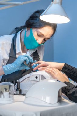 Salondaki bir kadın manikür ustası müşterinin tırnaklarıyla mavi arka planda çalışıyor. Kozmetik ve tırnak bakımı