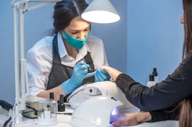 Salondaki bir kadın manikür ustası müşterinin tırnaklarıyla mavi arka planda çalışıyor. Kozmetik ve tırnak bakımı