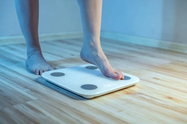 女人赤脚站在地板上的电子秤上 检查身体的重量 并在蓝光下控制体重的增加 — 图库照片
