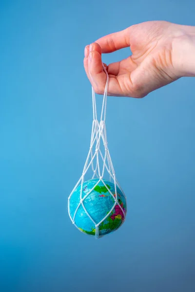 Hand holding a string bag with the planet Earth inside on a blue background. Concept: caring for nature, eco-friendly behavior, using reusable bags, fighting plastic