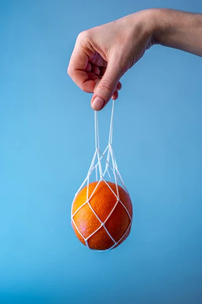 Hand holding a string bag with an orange on a blue background. Concept: caring for nature, eco-friendly behavior, fashion for using reusable bags