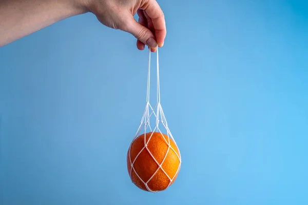 Hand holding a string bag with an orange on a blue background. Concept: caring for nature, eco-friendly behavior, fashion for using reusable bags