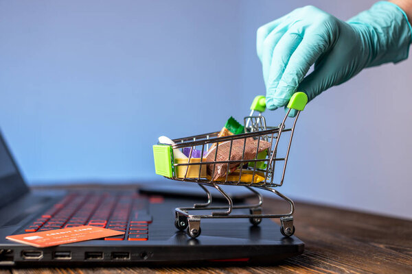 A hand in a sterile medical glove holds a shopping cart with a credit card for online purchases. Concept of internet purchasing important life support products during the coronavirus pandemic