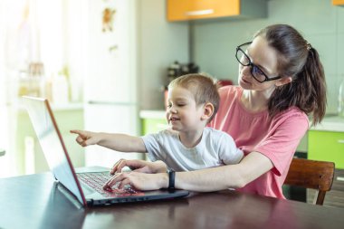 Dizlerinin üstünde küçük bir çocuğu olan genç bir kadın dizüstü bilgisayarın başında oturuyor. Uzaktan çalışma kavramı ve ev konforundan online eğitim alma.