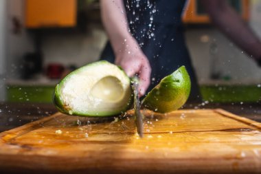 Şef hareket halindeki büyük bir bıçakla yeşil bir avokadoyu ikiye bölüyor. Su ve meyve suyu damlaları farklı yönlere uçar ve havada donar.