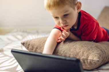 Anaokulundaki küçük bir çocuk, yerde bir yastığın üzerinde yatarken internetten tablet üzerinde çizgi film izliyor. Çocuklar için araçlar ve modern eğlence.
