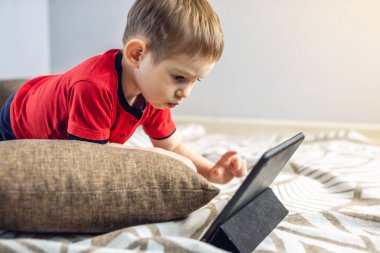 Anaokulundaki küçük bir çocuk, yerde bir yastığın üzerinde yatarken internetten tablet üzerinde çizgi film izliyor. Çocuklar için araçlar ve modern eğlence.