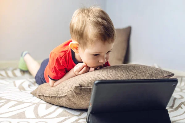 A preschool toddler child watches cartoons on a tablet on the internet while lying on a pillow on the floor. Gadgets and modern entertainment for children.