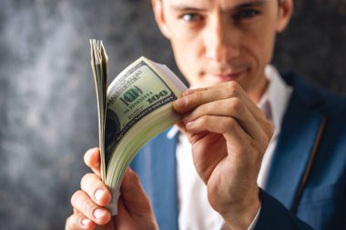 Takım elbiseli bir işadamı elinde bir yığın 100 dolarlık banknot tutuyor, gösterişli bir şekilde gösteriyor. Büyük miktarda para kazanma ve elde etme kavramı
