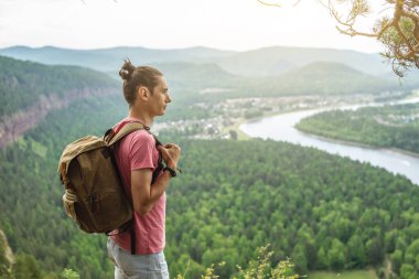 Sırt çantalı bir turist uçurumun kenarında duruyor ve nehri olan yeşil bir vadiye bakıyor. Özgürlük ve doğa ile birlik kavramı.