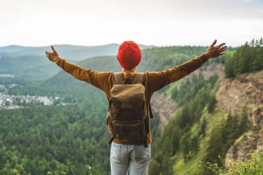Sırt çantası ve kırmızı şapkalı bir turist, yeşil bir vadinin önündeki uçurumun kenarında kollarını ardına kadar açmış duruyor. Özgürlük kavramı ve başarının sevinci.