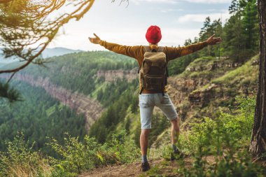 Sırt çantası ve kırmızı şapkalı bir turist, yeşil bir vadinin önündeki uçurumun kenarında kollarını ardına kadar açmış duruyor. Özgürlük kavramı ve başarının sevinci.